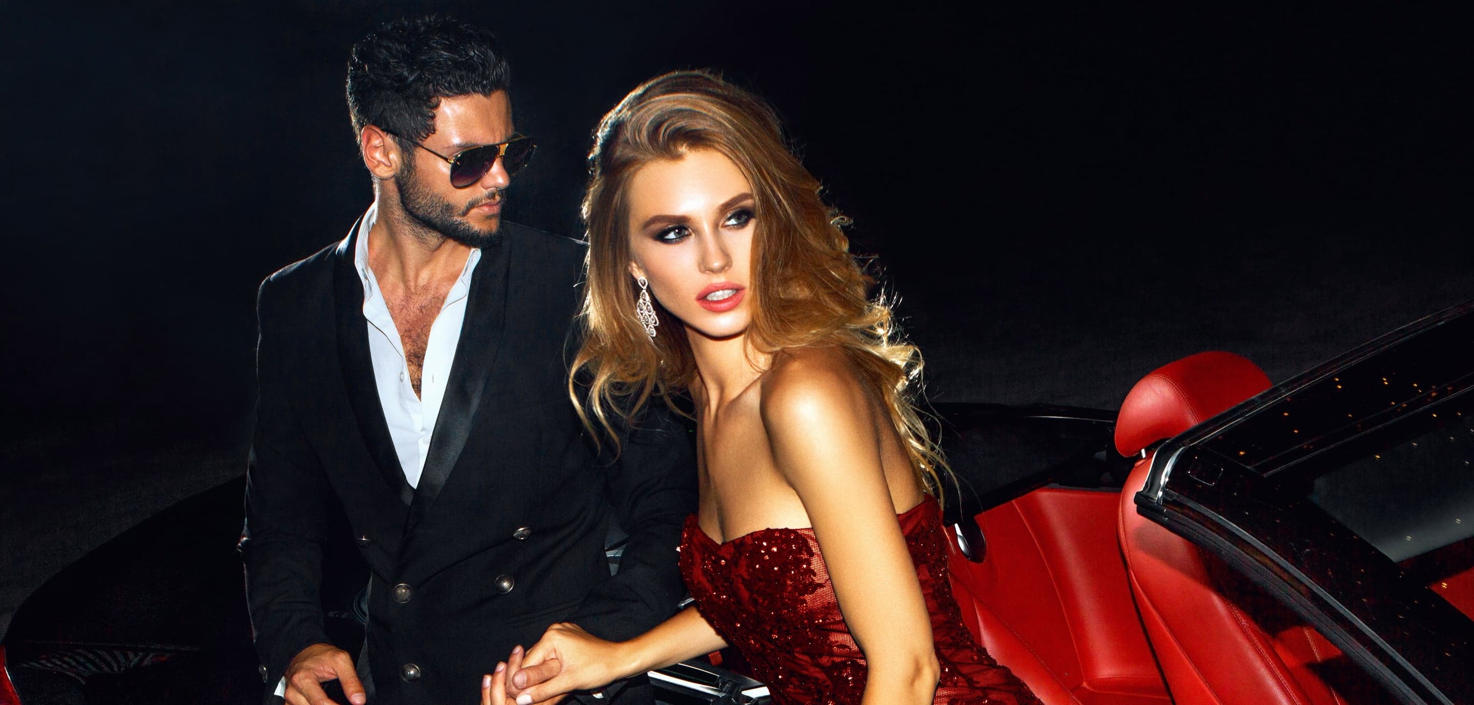 A man in a black tuxedo and a woman in a red sequinned dress beside a red convertible at night